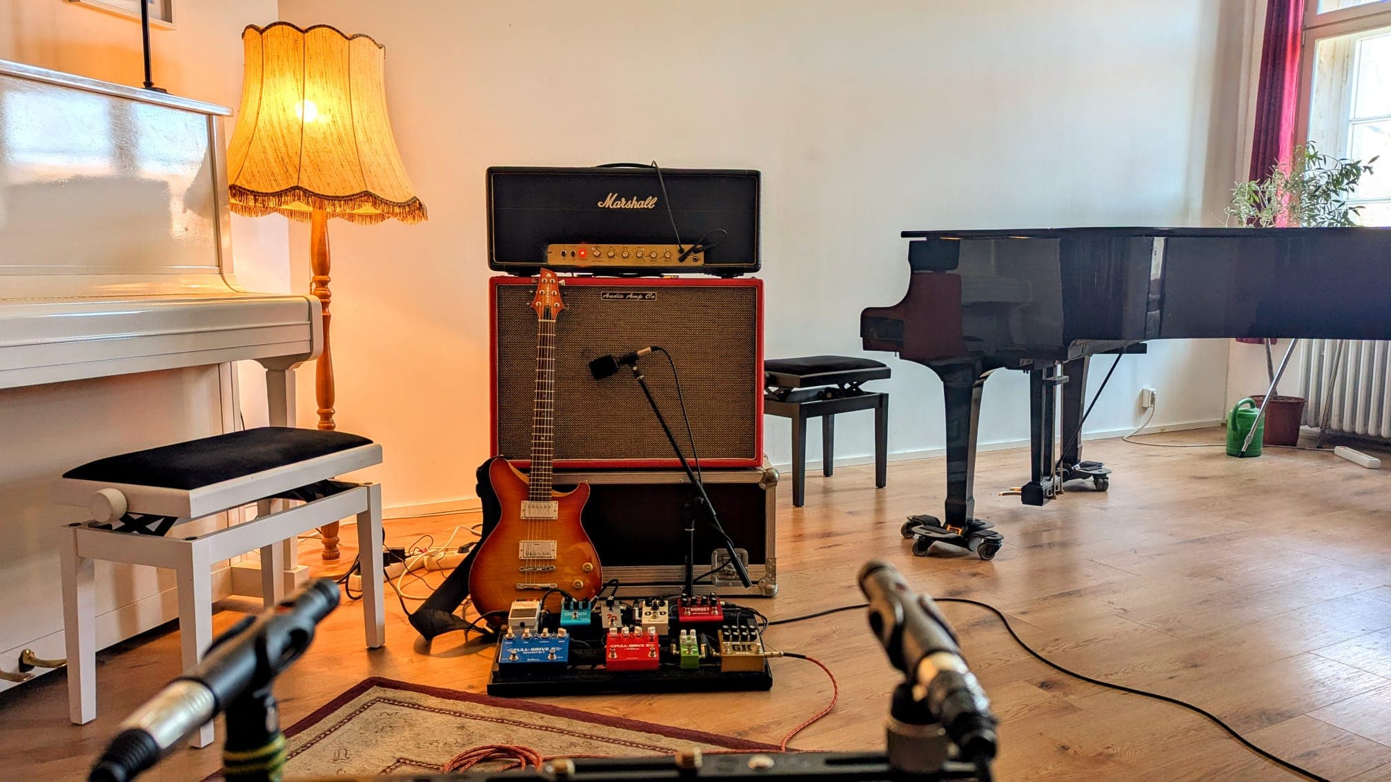 Ein helles Musikzimmer mit weißem Klavier, schwarzem Flügel, Marshall-Verstärker, E-Gitarre und Effektpedalen auf Holzboden.