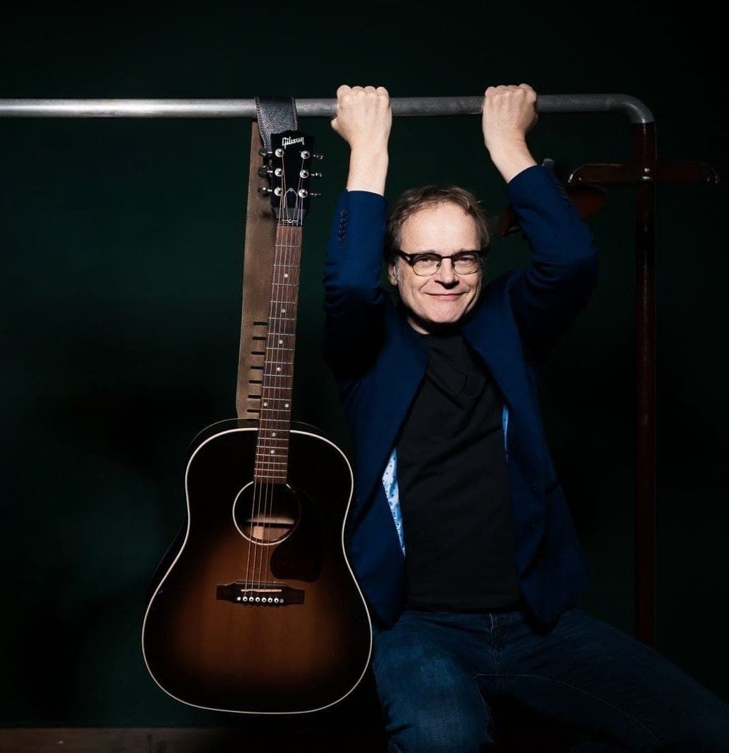 Ein Mann mit Brille hängt neben einer Gibson-Akustikgitarre an einer Metallstange vor dunklem Hintergrund.
