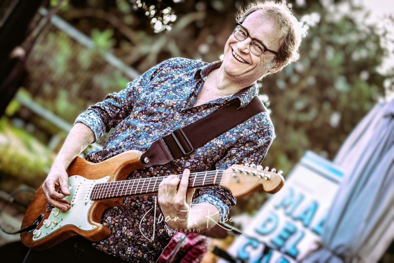 Lächelnder Mann mit Brille spielt eine hölzerne E-Gitarre im Freien und trägt ein geblümtes Hemd.
