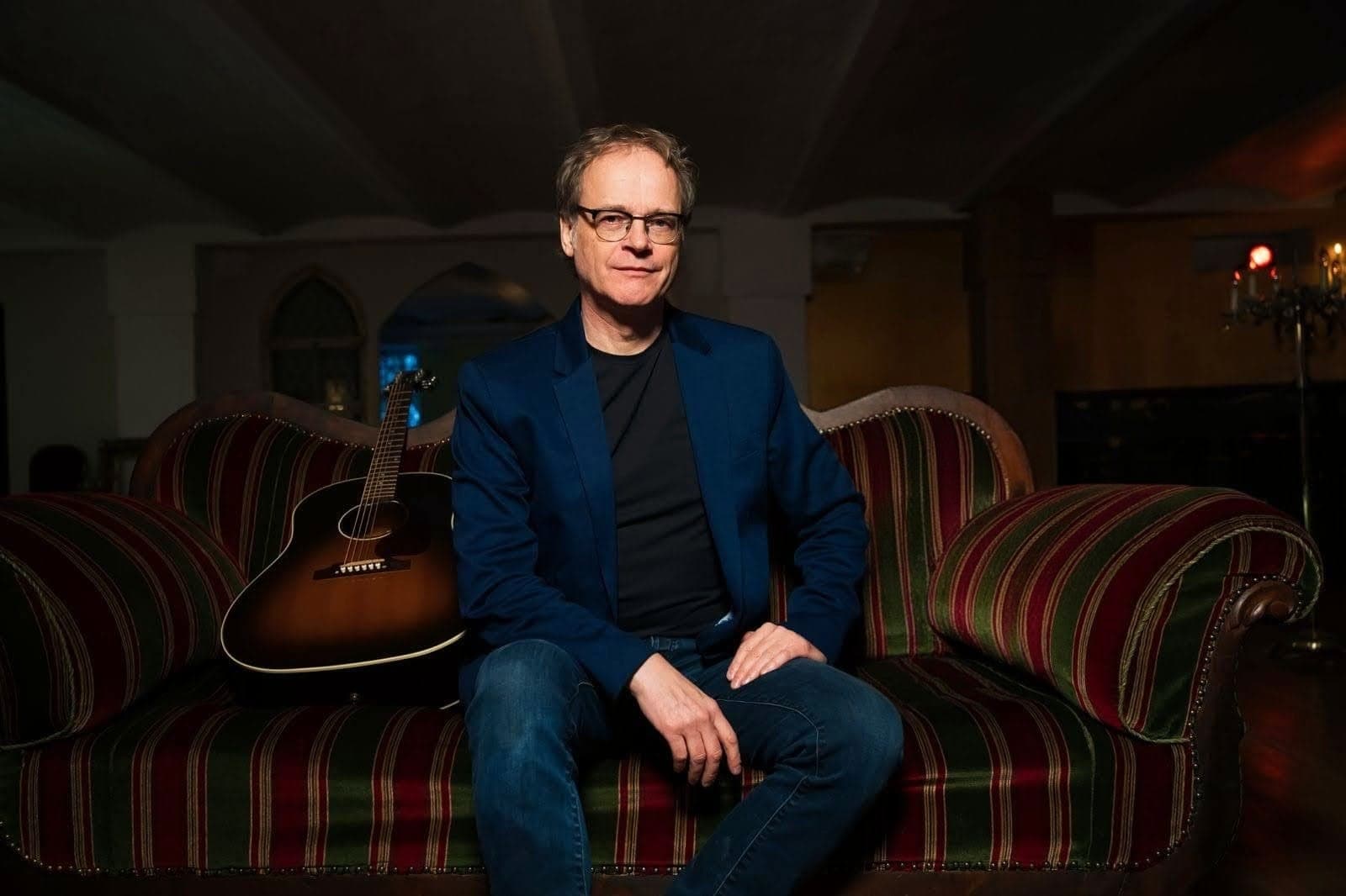 Mann mit Brille im blauen Sakko sitzt auf einem gestreiften Sofa neben einer Akustikgitarre.