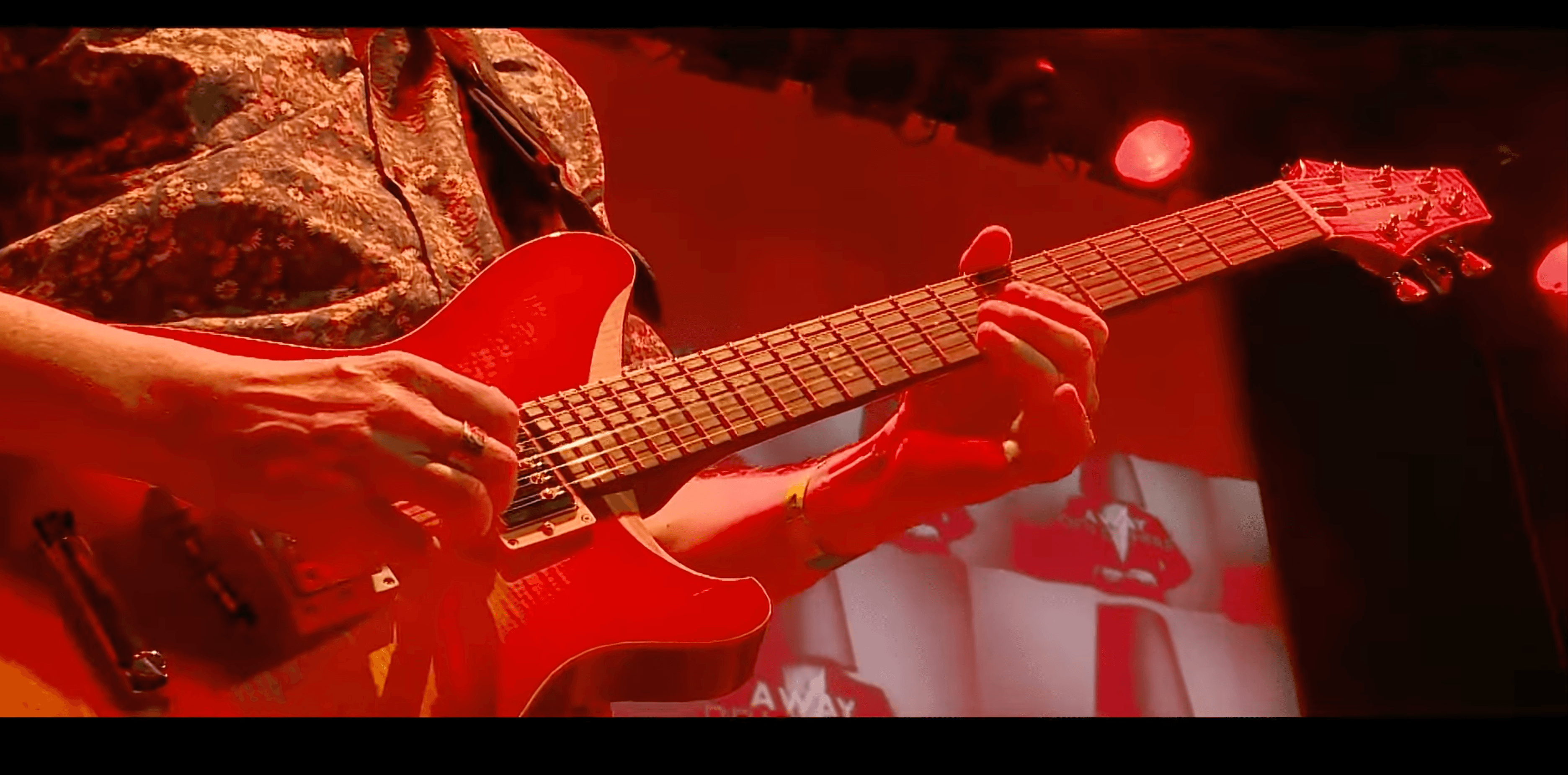 Nahaufnahme eines Gitarristen, der unter intensivem rotem Bühnenlicht auf einer elektrischen Gitarre spielt.