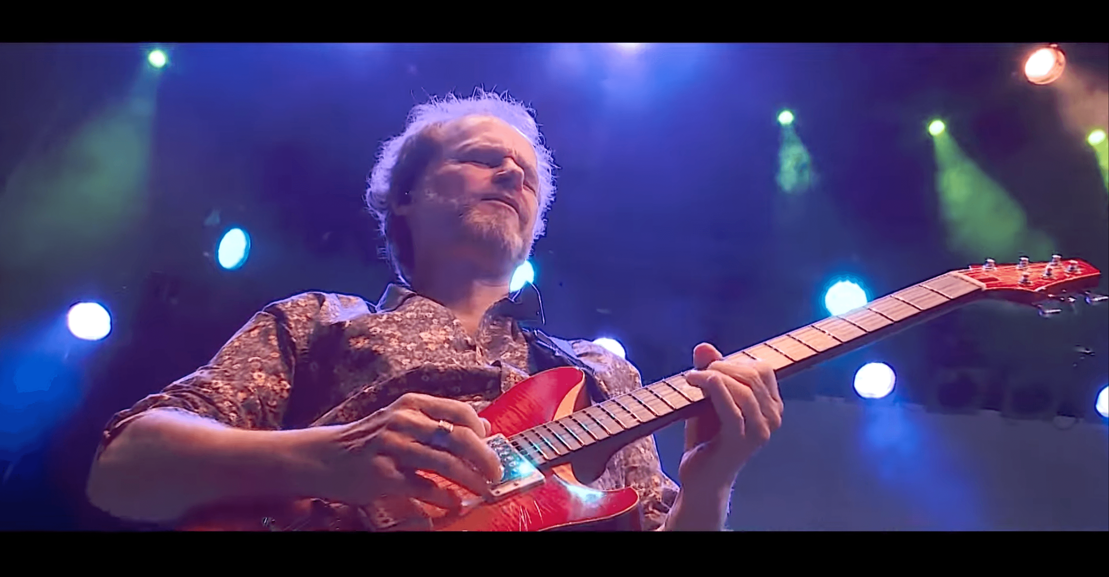 Bärtiger Musiker spielt mit geschlossenen Augen rote E-Gitarre auf einer Bühne unter blauem Scheinwerferlicht.
