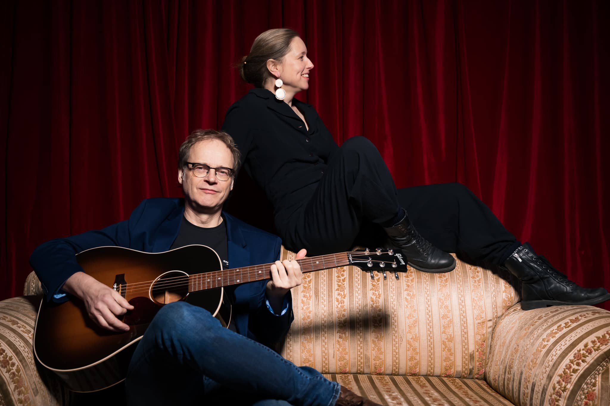 Ein Mann spielt Akustikgitarre neben einer Frau auf einem Vintage-Sofa vor roten Vorhängen.